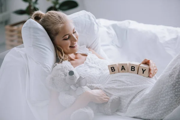 Боковой вид на улыбающуюся беременную женщину в белой ночнушке с плюшевым мишкой и деревянными блоками с детскими надписями на животе, лежащих на диване — стоковое фото
