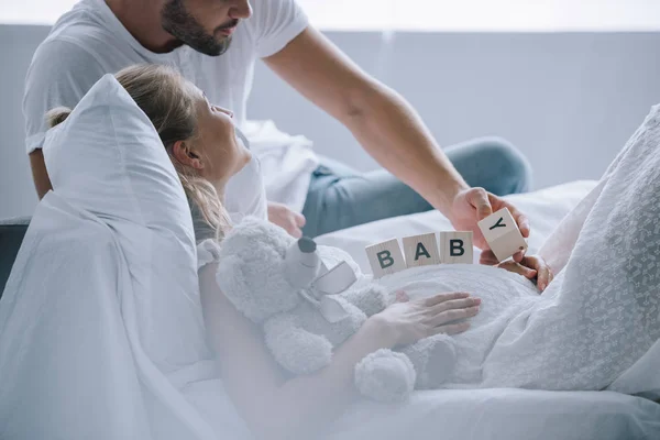 Partial view of man putting wooden blocks with baby lettering on belly of pregnant wife with teddy bear at home — Stock Photo
