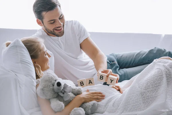 Homem sorrindo colocando blocos de madeira com letras de bebê na barriga da esposa grávida com ursinho de pelúcia em casa — Fotografia de Stock