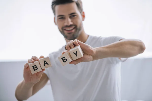 Retrato de homem sorridente mostrando blocos de madeira com inscrição de bebê em mãos em casa — Fotografia de Stock