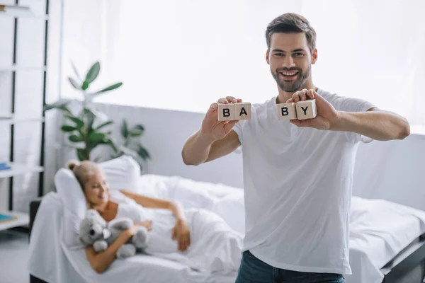 Foco seletivo de homem sorridente mostrando blocos de madeira com inscrição do bebê enquanto a mulher grávida descansando no sofá — Fotografia de Stock