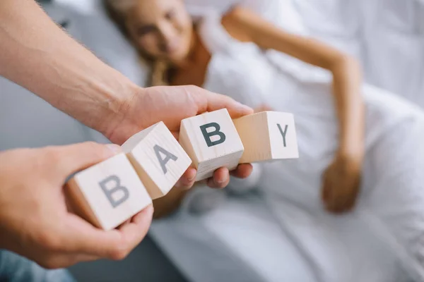 Ritagliato colpo di uomo che tiene blocchi di legno con scritte per bambini mentre la moglie incinta riposa sul divano a casa — Foto stock