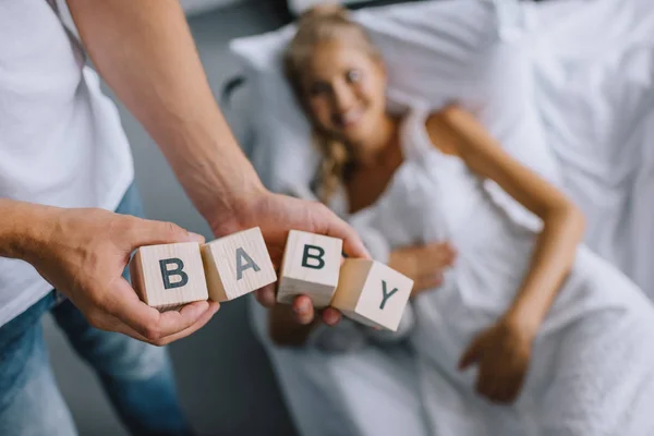 Cropped shot of man holding wooden blocks with baby lettering while pregnant wife resting on sofa at home — Stock Photo