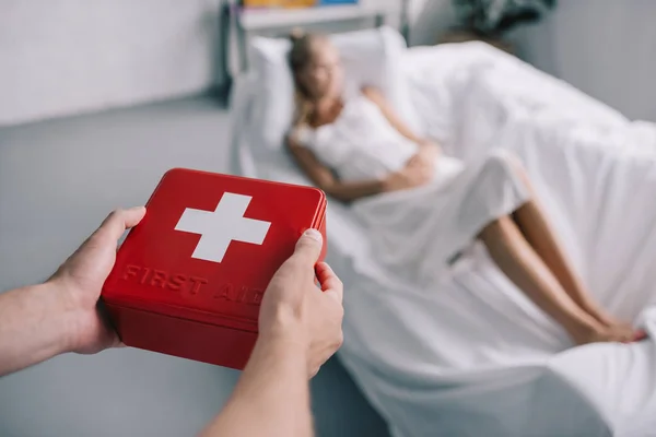 Cropped shot of man bringing first aid kit to pregnant wife in white nightie on sofa at home — Stock Photo