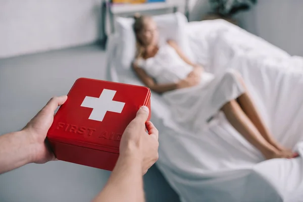 Tiro cortado do homem trazendo kit de primeiros socorros para a esposa grávida em camisa de noite branca no sofá em casa — Fotografia de Stock