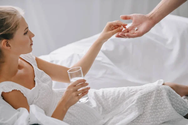 Partial view of man giving medicines to pregnant woman in white nightie on sofa at home — Stock Photo