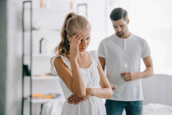 Pregnant woman in white nightie having headache while husband with medicines and glass of water standing behind at home — Stock Photo