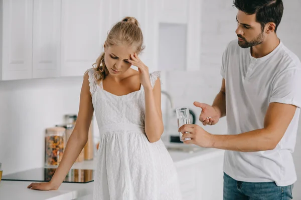 Pregnant woman in white nightie having headache while husband giving medicines and glass of water to her at home — Stock Photo