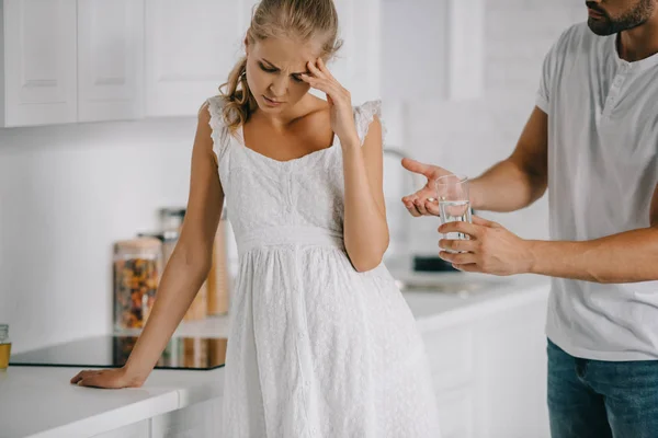 Schwangere im weißen Nachthemd hat Kopfschmerzen, während ihr Mann ihr zu Hause Medikamente und ein Glas Wasser gibt — Stockfoto