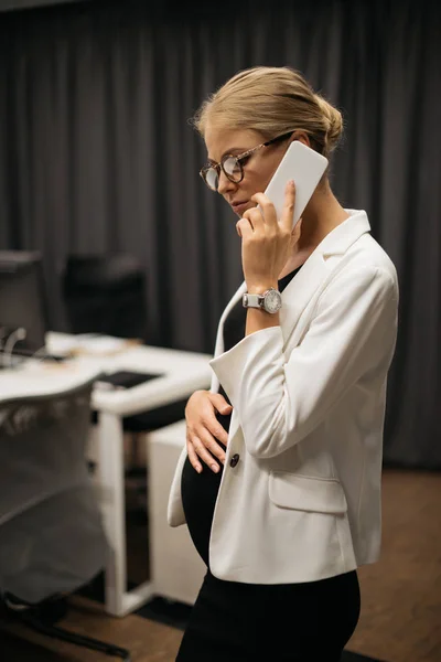 Vista lateral de mujer de negocios embarazada en ropa formal hablando en el teléfono inteligente en la oficina - foto de stock