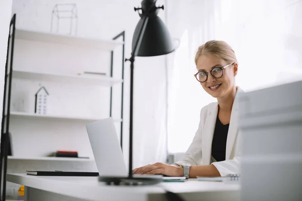 Портрет усміхнений бізнес-леді в окулярів, дивлячись на камеру на робочому місці з ноутбуком в офісі — стокове фото