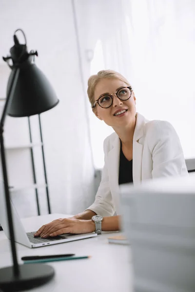 Retrato de mujer de negocios en gafas mirando hacia el lugar de trabajo con el ordenador portátil en la oficina - foto de stock