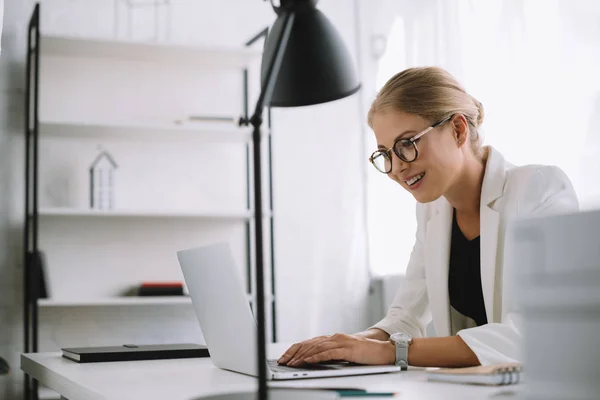 Вид сбоку улыбающейся деловой женщины, работающей на ноутбуке на рабочем месте в офисе — стоковое фото