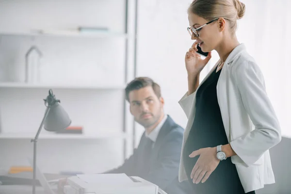 Lächelnde schwangere Geschäftsfrau spricht auf Smartphone, während sie neben Kollegin am Arbeitsplatz im Büro steht — Stockfoto