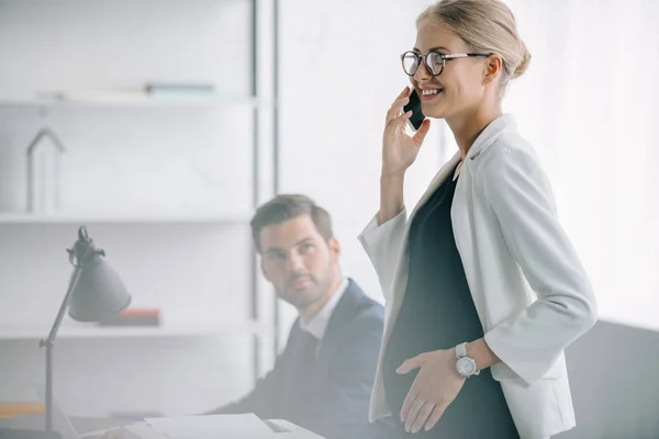 Smiling pregnant businesswoman talking on smartphone while standing near colleague at workplace in office — Stock Photo