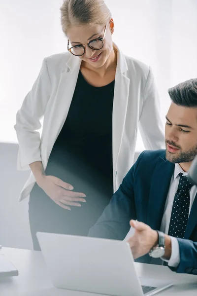 Portrait of pregnant businesswoman and colleague at workplace with laptop discussing project in office — Stock Photo