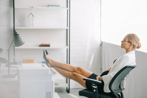 Vista lateral de la mujer de negocios embarazada descansando con las piernas en la mesa en el lugar de trabajo en la oficina - foto de stock