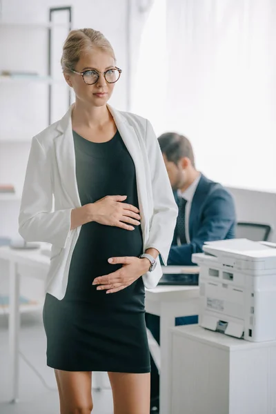 Selective focus of pensive pregnant businesswoman and colleague at workplace behind in office — Stock Photo