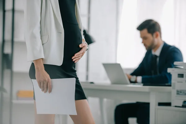 Partial view of pregnant businesswoman with documents and colleague working on laptop at workplace in office — Stock Photo