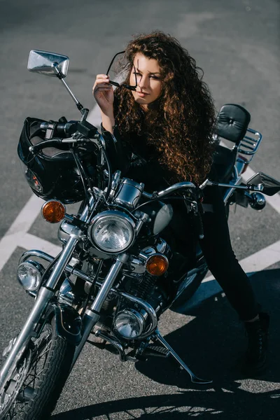 Кучерява жінка сидить на подрібненому мотоциклі з шоломом на міській парковці — стокове фото