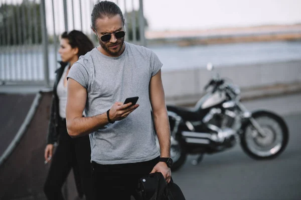 Homme en lunettes de soleil en utilisant smartphone tandis que la petite amie debout avec moto classique sur fond — Photo de stock