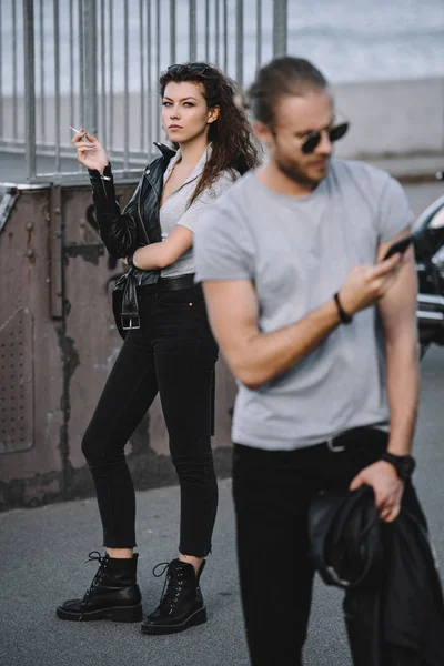 Novio usando teléfono inteligente, mientras que la novia de pie con la motocicleta clásica en el fondo - foto de stock