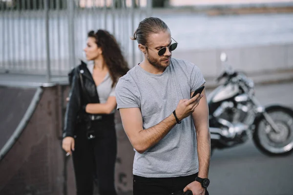 Man using smartphone while girl standing with classical motorbike on background — Stock Photo