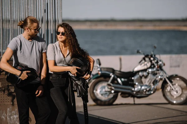 Pareja de jóvenes ciclistas sosteniendo cascos, motocicleta clásica en el fondo - foto de stock