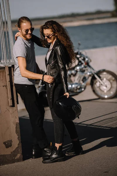 Young couple of bikers embracing in city with motorbike on background — Stock Photo