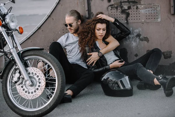 Пара байкеров, обнимающихся и сидящих на асфальте со шлемом и мотоциклом — стоковое фото