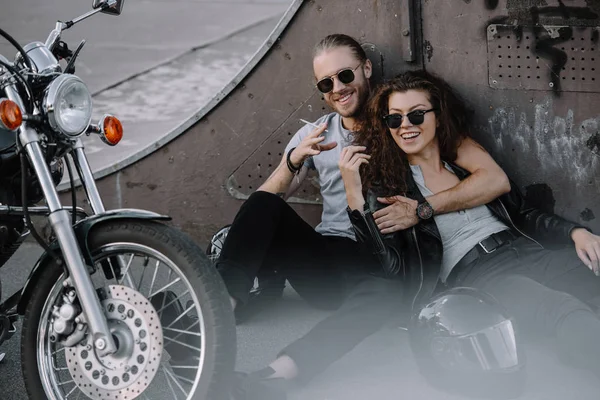 Sorrindo casal de motociclistas abraçando e fumando no asfalto com motocicleta helicóptero — Fotografia de Stock