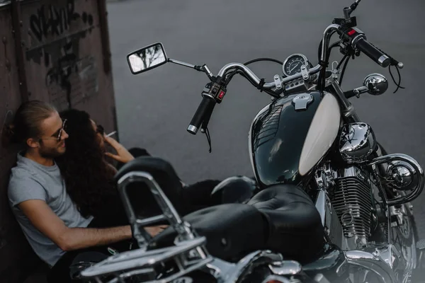Foco seletivo de motociclistas fumando e sentado no asfalto perto de motocicleta clássica — Fotografia de Stock
