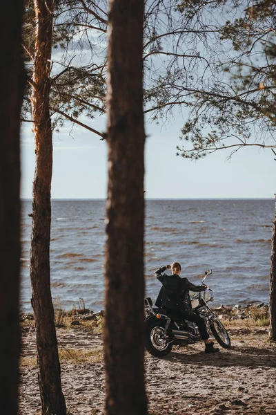 Байкер в черной кожаной куртке сидит на классическом мотоцикле вертолета возле реки в лесу — стоковое фото