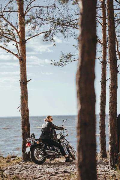 Biker in black leather jacket sitting on classic motorcycle near the sea in forest — Stock Photo