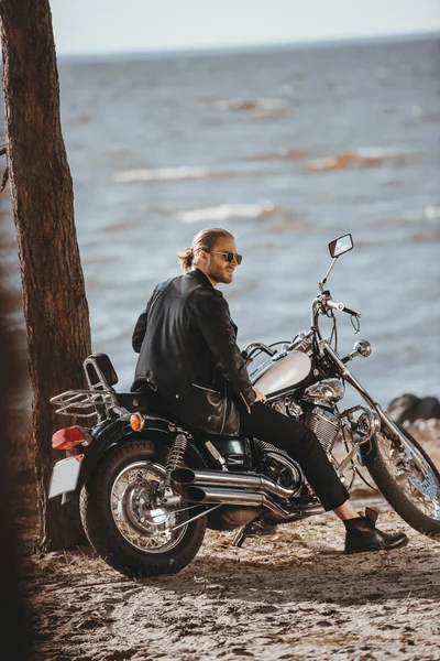 Motociclista em jaqueta de couro preto sentado na motocicleta helicóptero clássico perto do mar — Fotografia de Stock