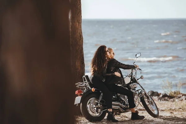 Пара в черных кожаных куртках сидит на винтажном мотоцикле и смотрит на море — стоковое фото