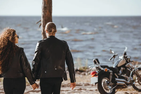 Vista posteriore della coppia che si tiene per mano e cammina sulla riva del mare, moto classica in piedi vicino — Foto stock