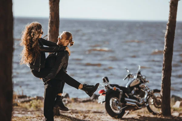 Щасливий чоловік тримає свою дівчину на руках на березі моря з класичним мотоциклом поруч — стокове фото