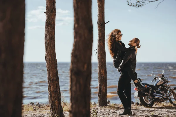 Copain tenant sa petite amie heureuse sur les bras sur le bord de la mer avec moto près — Photo de stock