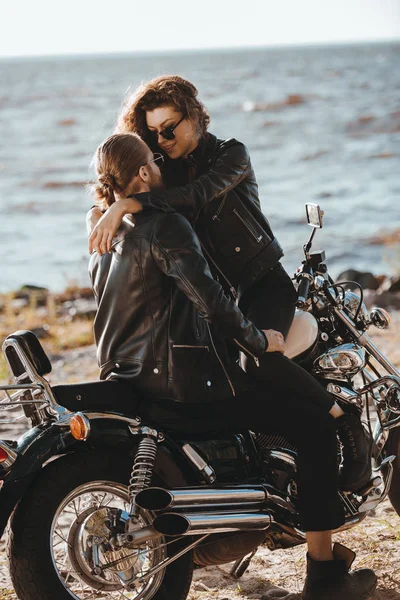 Pareja en chaquetas de cuero negro abrazándose en moto vintage en la orilla del mar - foto de stock