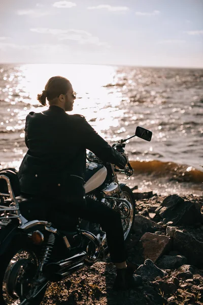 Motero guapo sentado en motocicleta clásica cerca del mar con luz de fondo - foto de stock