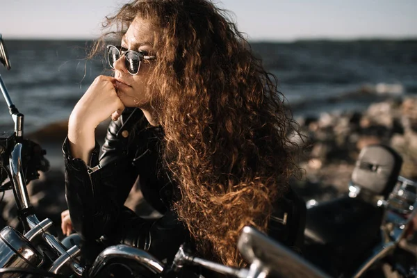 Привлекательная кудрявая байкер в солнечных очках, сидящая на мотоцикле — стоковое фото