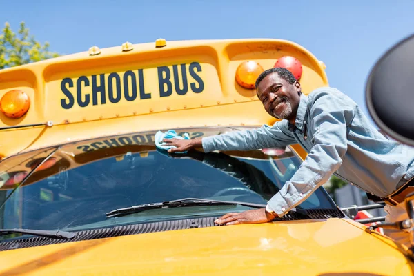 Sonriente maduro afroamericano autobús conductor limpiando la ventana delantera de autobús escolar y mirando a la cámara - foto de stock