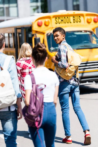 Vista posteriore di bello teen africano americano scolaro a piedi a scuolabus con i suoi compagni di classe e tornando alla fotocamera — Foto stock