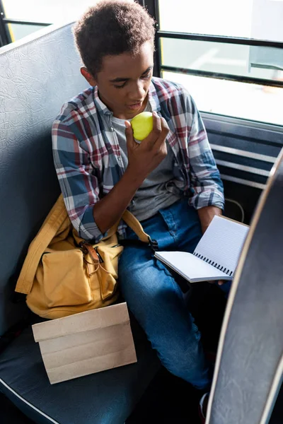 Американский школьник-подросток читает яблоко, держа в руках чистый блокнот. — стоковое фото