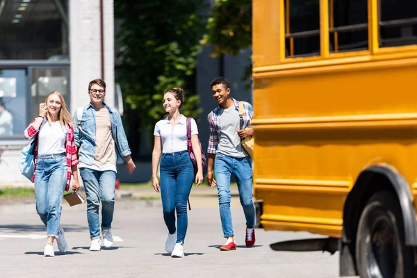 Grupo de adolescentes eruditos caminando detrás del autobús escolar en el estacionamiento - foto de stock
