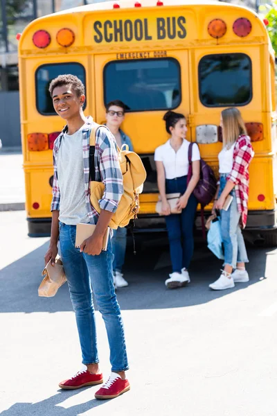 Красивый улыбающийся школьник смотрит в камеру, в то время как его одноклассники стоят позади школьного автобуса — стоковое фото