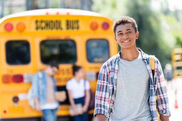 Glücklich teen afrikanisch amerikanisch schoolboy looking at camera vor school bus — Stockfoto