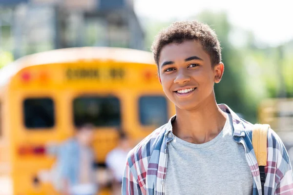 Heureux adolescent afro-américain écolier regardant caméra avec flou autobus scolaire sur fond — Photo de stock