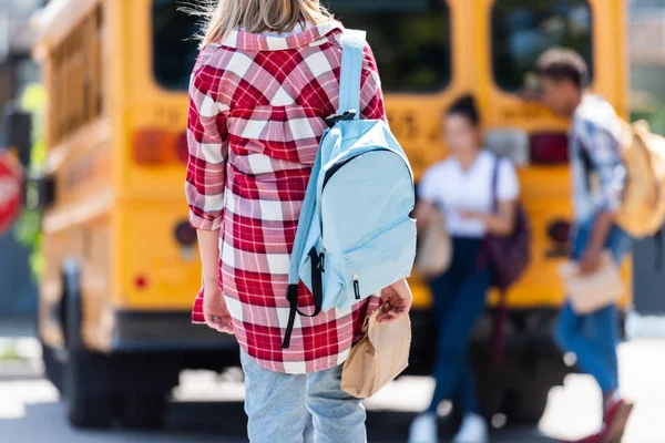 Rear view of teen schoolgirl walking to classmates leaning on school bus — Stock Photo